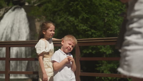 bored little children stand on bridge near waterfall in wild park. boy and girl tired of long walk to cascade on mountain river on summer vacation