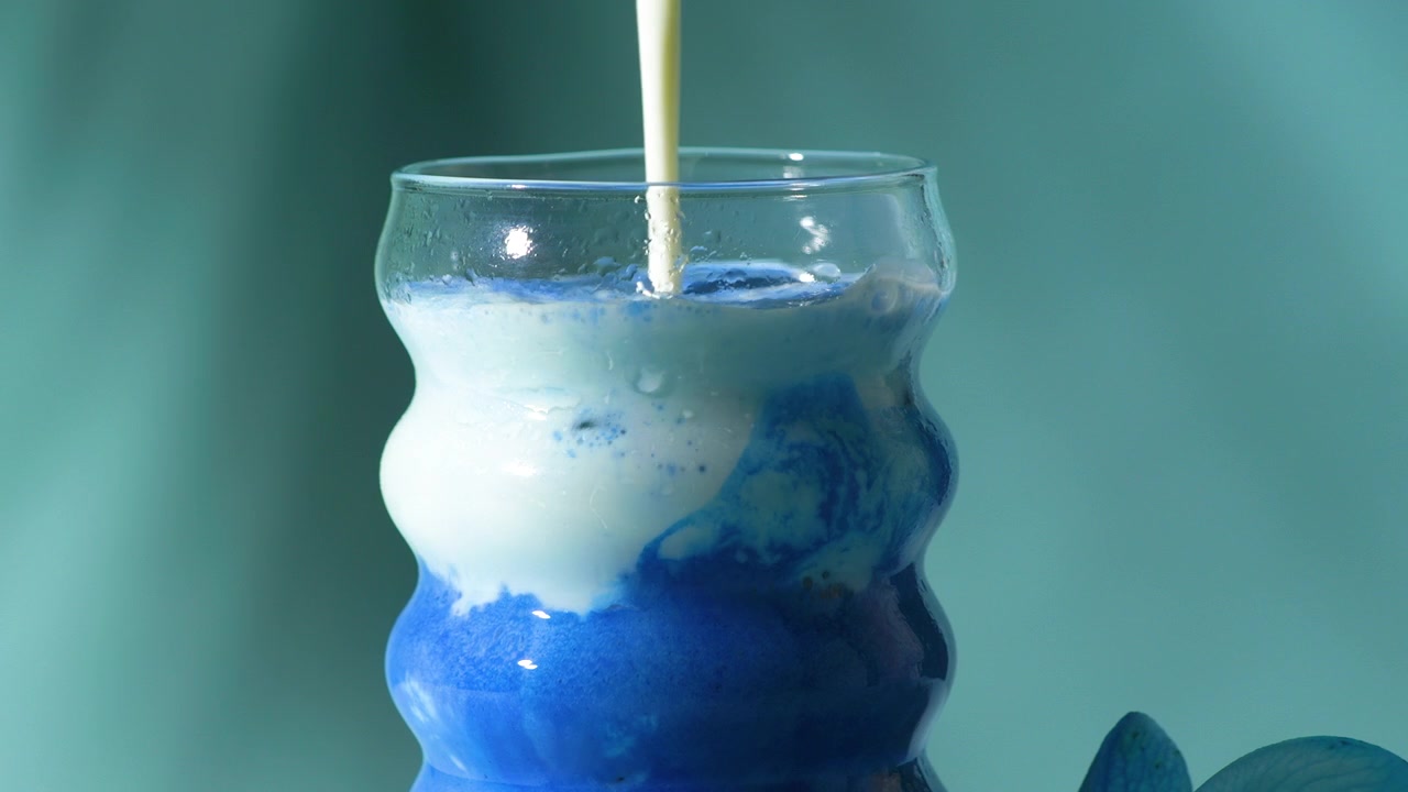 Melk helles sakte i en levende blå sommerfuglertedrikk i et bølget bobleglass, noe som skaper en virveleffekt på en turkis bakgrunn.