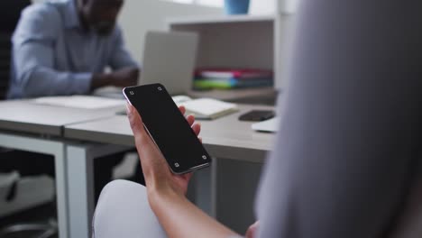 Mixed-race-businesswoman-sitting-at-desk-and-using-smartphone-with-copy-space-on-screen-in-office