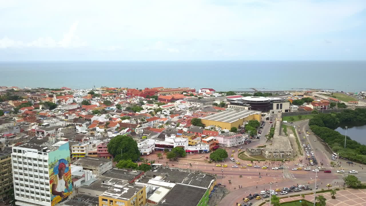 Aerial zoom in toward coastal city with dense urban buildings, harbor area and ocean horizon under bright daylight