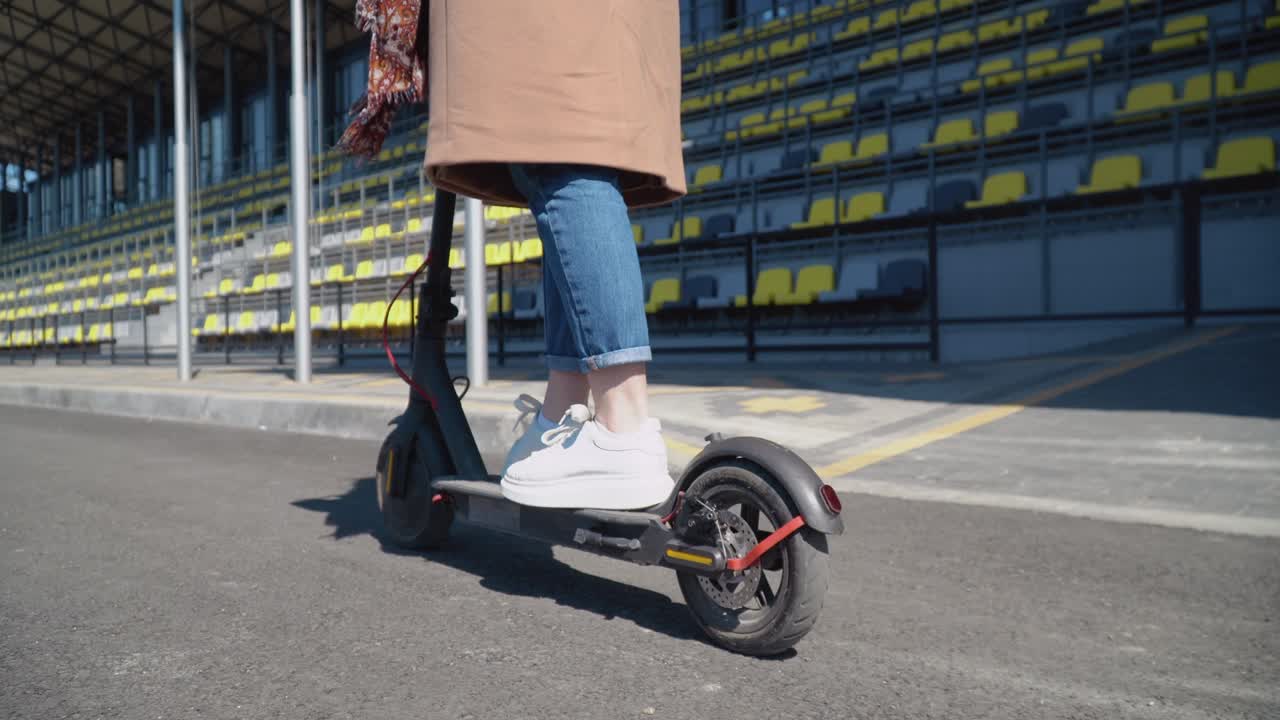 ジーンズとコートを着た女子学生が電動スクーターに乗っている。スクーターと車輪の後方からのクローズアップ。