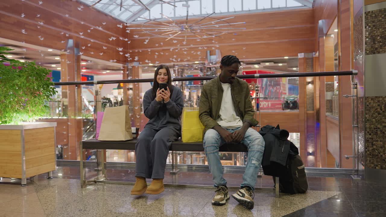 Lady using smartphone beside young man sitting on bench with shopping bags inside spacious modern mall decorated with wood panels, indoor plants, bird ornaments, and natural light from glass roof
