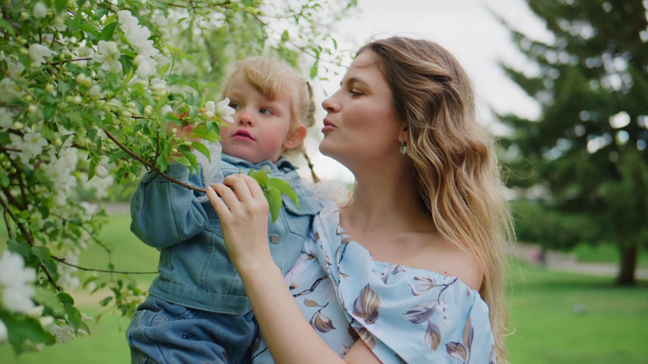 Mutter hält Kleinkind unter blühendem Baum. Kind greift nach Blüten, kichert fröhlich. Mutter lächelt warm und teilt einen friedlichen Moment. Sonnenlicht filtert durch weiße Blüten