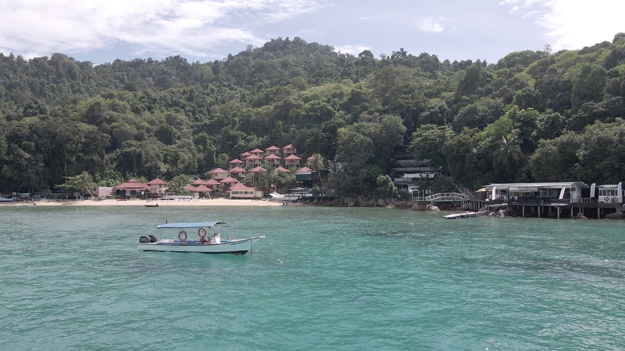 Aerial video of a boat floating on anchor in front of a remote island resort. Filmed at the Alunan resort, Perhentian Kecil, Malaysia. Slow dolly forward above the ocean.