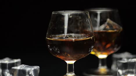 two glasses of brown liquor on a black background with ice cubes