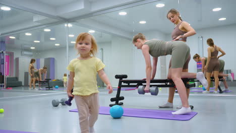 trainer places hand on client back, correcting posture during dumbbell row workout, focused trainee lifts weight while gym mirror reflects environment, child with small dumbbell walks toward camera