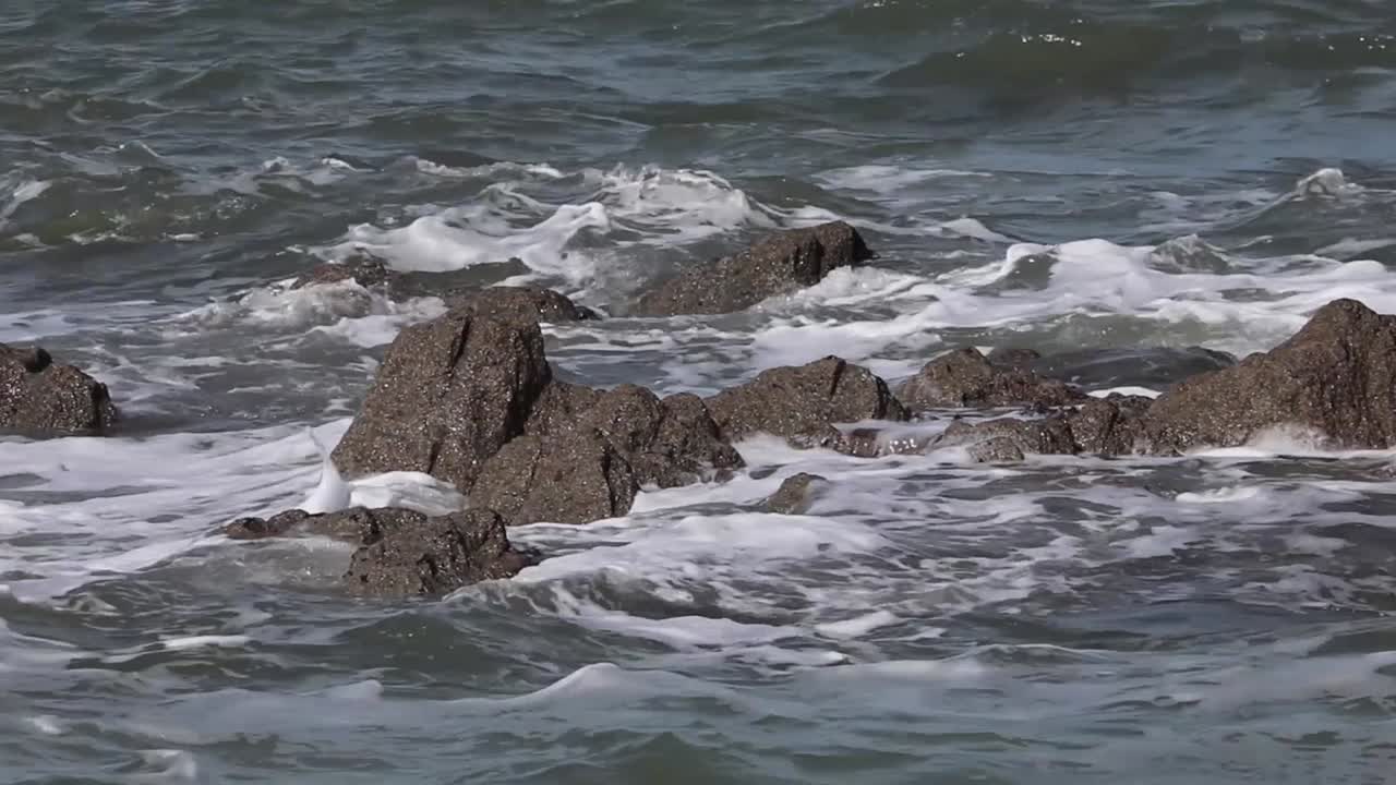 Ocean waves crashing against exposed rocks along the coast of Cornwall, England, showing white water splash, rough sea movement, and natural coastal power in daylight