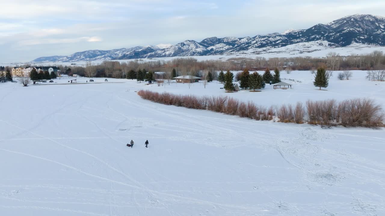 Boseman Montana Aerial Winter sunset ice fishermen pulling sled over snowy suburban park, orbit right with 4k drone with mountain backdrop at golden hour