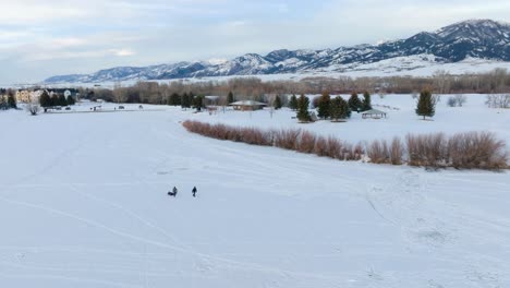 Boseman-Montana-Antena-Invierno-Puesta-De-Sol-Pescadores-De-Hielo-Tirando-Del-Trineo-Sobre-El-Parque-Suburbano-Nevado,-Orbitan-A-La-Derecha-Con-Un-Dron-De-4k-Con-Fondo-De-Montaña-En-La-Hora-Dorada