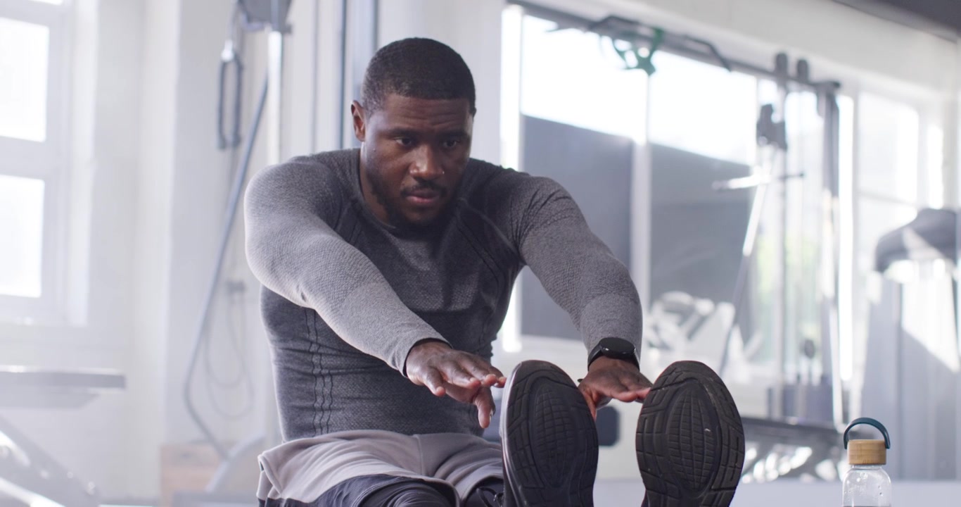 African male doing seated leg stretch at gym, reaching to shoes with watch, bottle during warmup