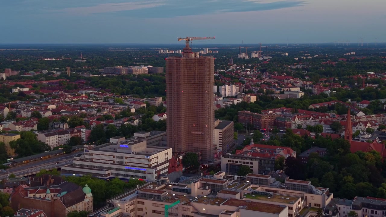 Skyskraber under opførelse i et byområde med en kran på toppen, omgivet af beboelsesejendomme og træer ved solnedgang. Fantastisk omvendt dronepanorama fra luften.
