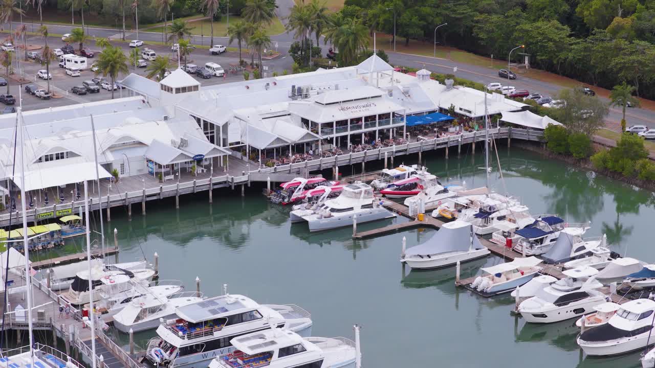 Aerial footage captures boats docked at Port Douglas marina, with surrounding buildings and lush greenery under soft daylight