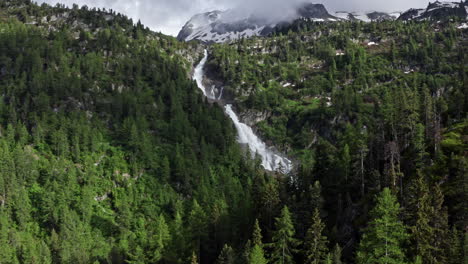 Una-Impresionante-Cascada-Cae-A-Través-De-Un-Exuberante-Bosque-Verde-Con-Montañas-Nevadas-Al-Fondo.