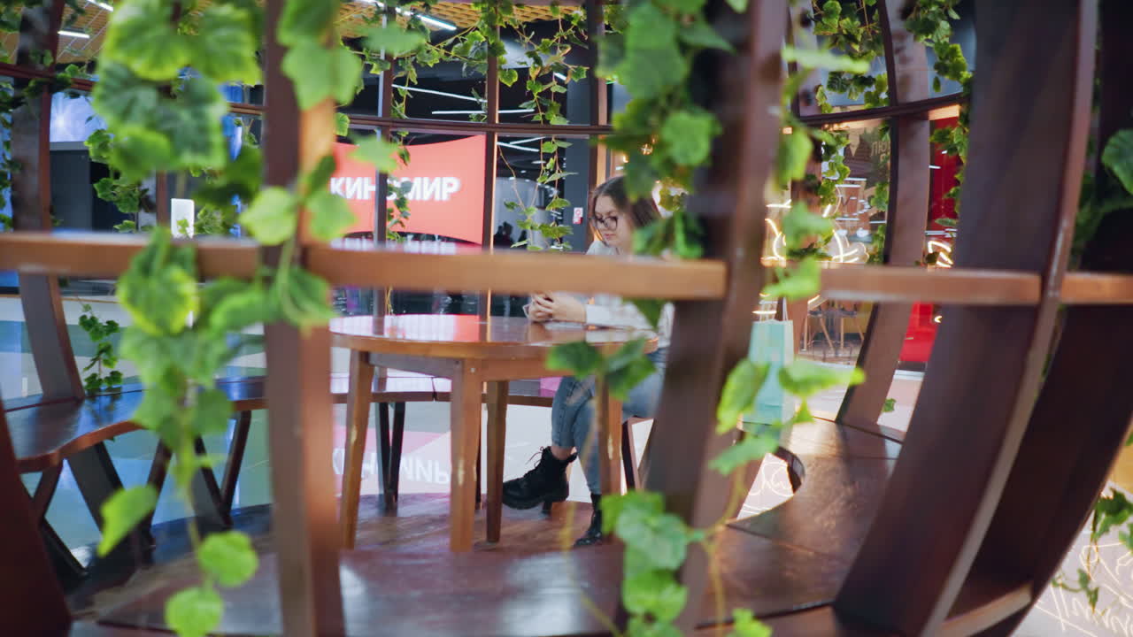 트렌디하고 편안한 분위기를 조성하는 초록색으로 둘러싸인 세련된 실내 바 영역에서 다리를 은 나무 테이블에 앉아있는 여인