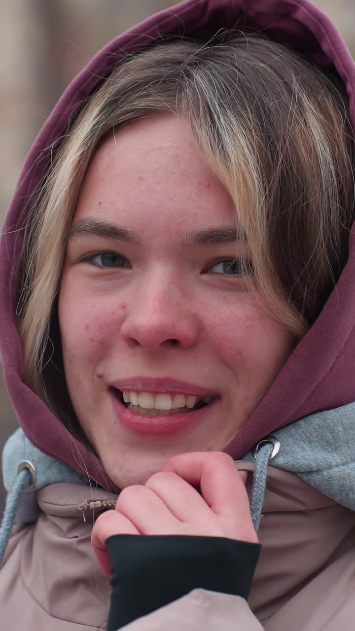 Shy young woman in hoodie gently holds hoodie collar while sitting on swing in winter park, dressed in warm jacket, she enjoys cold weather with a soft smile and blurred background featuring trees
