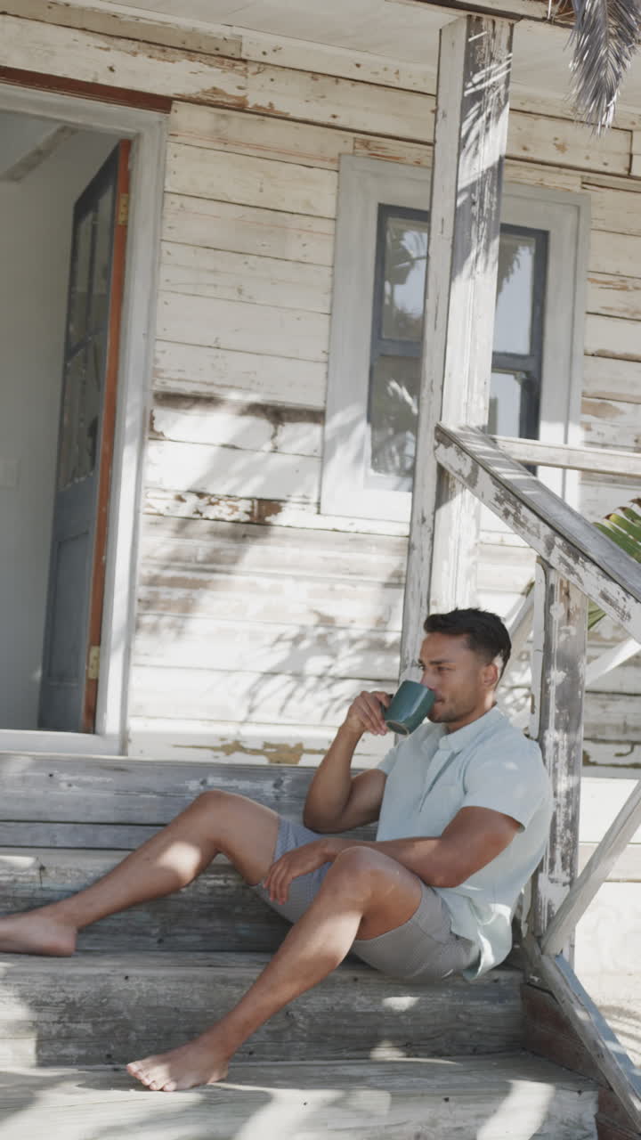 Vertical video of thoughtful biracial man drinking coffee on steps of beach house, slow motion