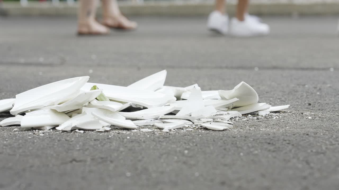 Shattered white plate pieces on asphalt with people walking in the background