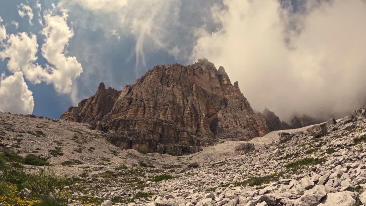 Timelapse video of a rugged mountain cliff rises sharply beneath a dramatic sky, partially shrouded in mist and clouds. The foreground is scattered with rocks and alpine vegetation.