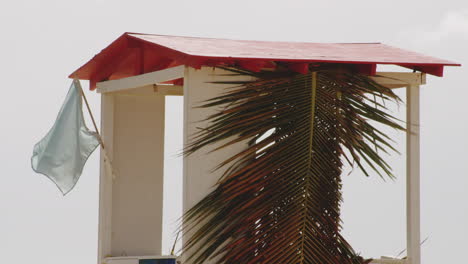 Flag-Blowing-In-The-Wind-At-Lifeguard-Shack-With-Coconut-Leaves-At-Balneario-del-Escambron,-Beach-In-San-Juan,-Puerto-Rico