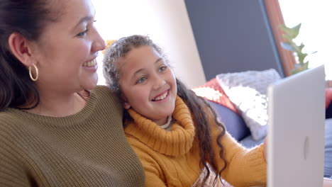 happy biracial mother and daughter sitting on sofa, using laptop and smiling in sunny living room