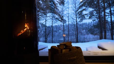 beautiful view of snow-covered trees in the forest and the lake through the window of a house with a burning fireplace in cold winter evening