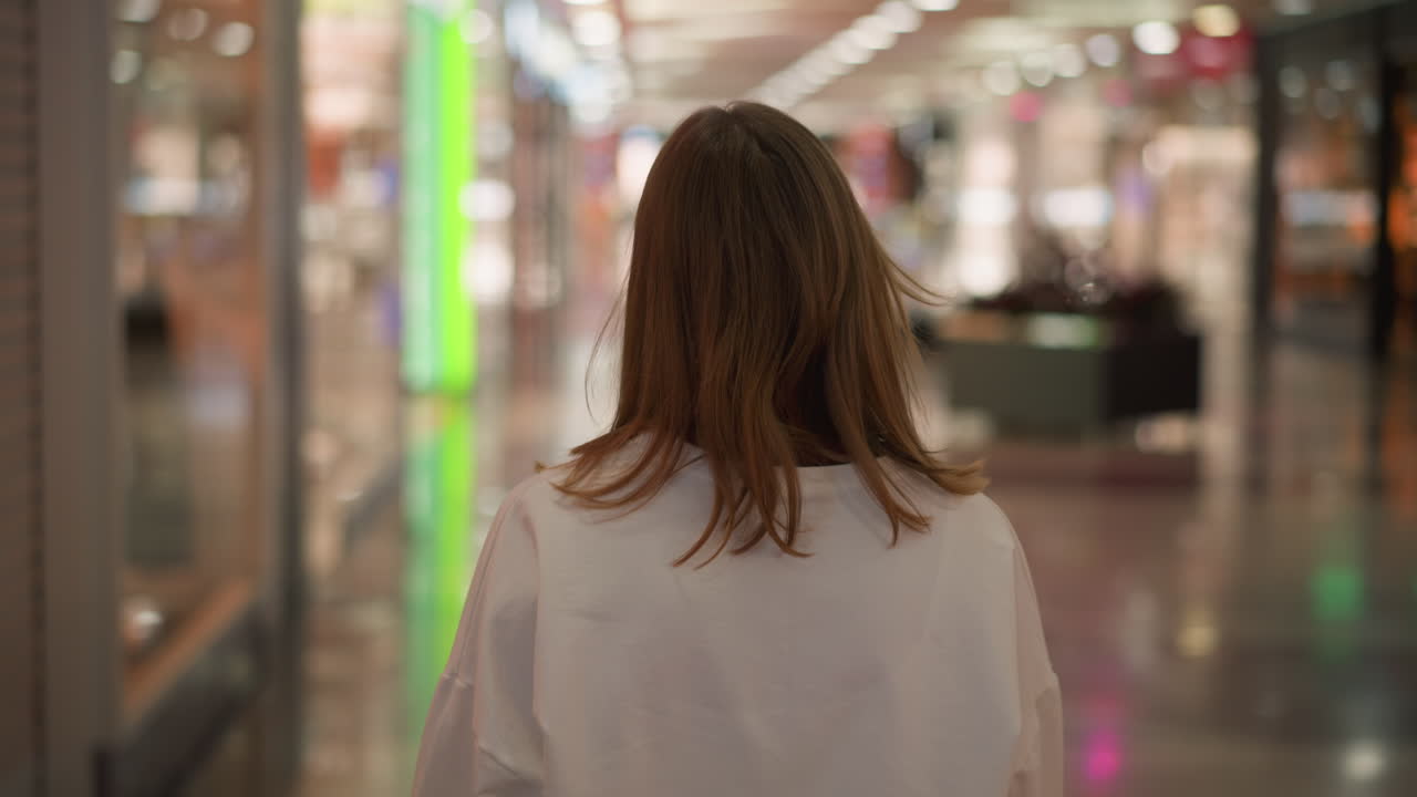 Back view of a woman in a white top walking through a shopping mall, illuminated by colorful lights with a blurred background featuring reflective glass surfaces