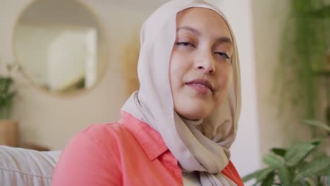 video of thoughtful biracial woman in hijab writing and looking away smiling in living room at home