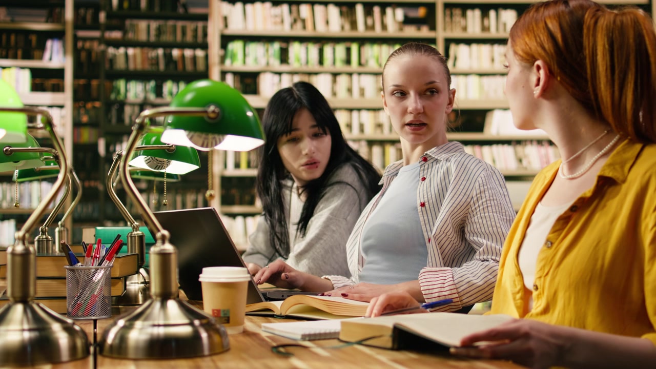 Studenti che lavorano fianco a fianco a un tavolo della biblioteca con laptop e libri aperti