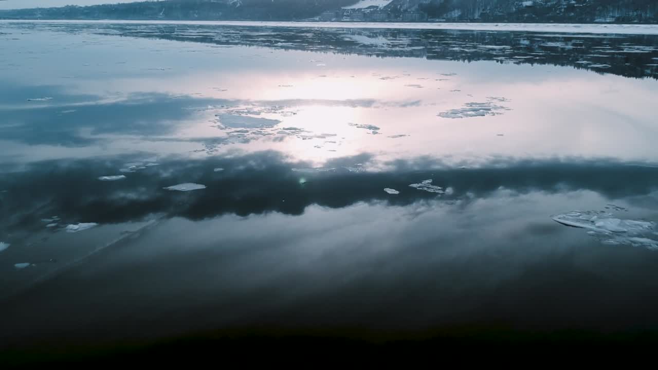 Sunlit ice floes drift across calm river surface during winter season as soft light reflects on frozen water creating peaceful natural scene captured from drone perspective