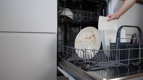 man opening a dishwasher and loading it with dirty plates and cups before closing it.