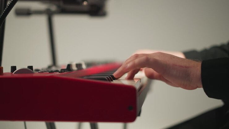 A keyboardist in a black outfit sits and plays a red sampler against a grey background. The focus is on the hands playing the keyboard, highlighting the detailed and skilled performance