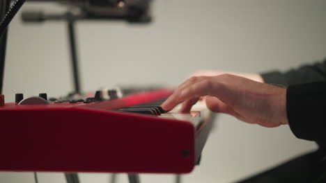 a keyboardist in a black outfit sits and plays a red sampler against a grey background. the focus is on the hands playing the keyboard, highlighting the detailed and skilled performance