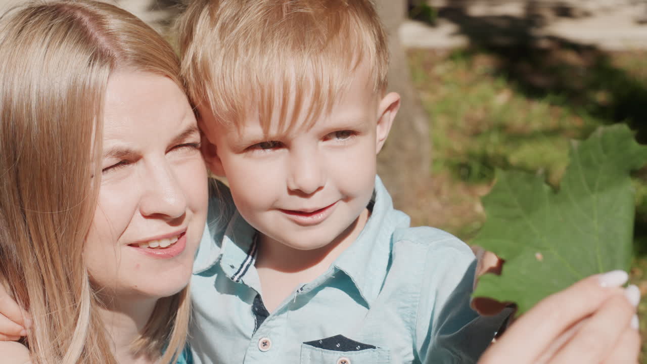Madre e figlio nel parco durante il weekend, tengono una foglia e sorridono sotto la calda luce del sole. Un momento familiare casuale all'aperto con un abbraccio affettuoso, alberi verdi e una scoperta giocosa. Un ritratto estivo spontaneo che cattura la gioia.