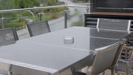 static shot of a wet balcony at a hotel during steady rain, with water droplets on glass railing and a reflective metal table surface