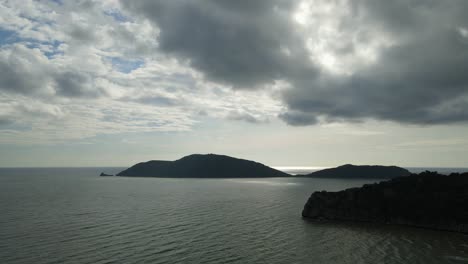 A-steady-aerial-footage-of-an-island-in-the-horizon-and-a-mountain-on-the-right-hand-side,-heavy-rain-clouds-and-blue-sky