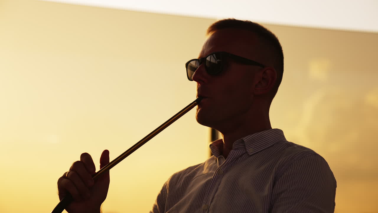 Man in dark glasses sitting at sunset holding hookah pipe. Relaxing male making thick smoke from nargileh. Close up.