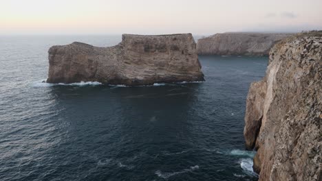 establishing shot, scenic view of a wave hitting the rock formation near the shoreline of algarve, portugal, cloudy misty sky in the background