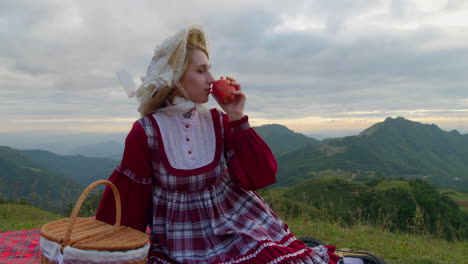 Medium-view-of-woman-picking-up-apple-from-red-picnic-blanket-overlooking-mountain-vista