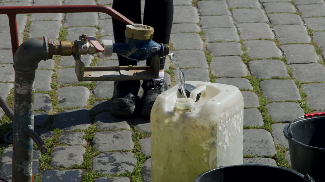 Persona riempie un grande contenitore d'acqua di plastica da un rubinetto pubblico in strada alla luce del giorno, con sfondo di ciottoli