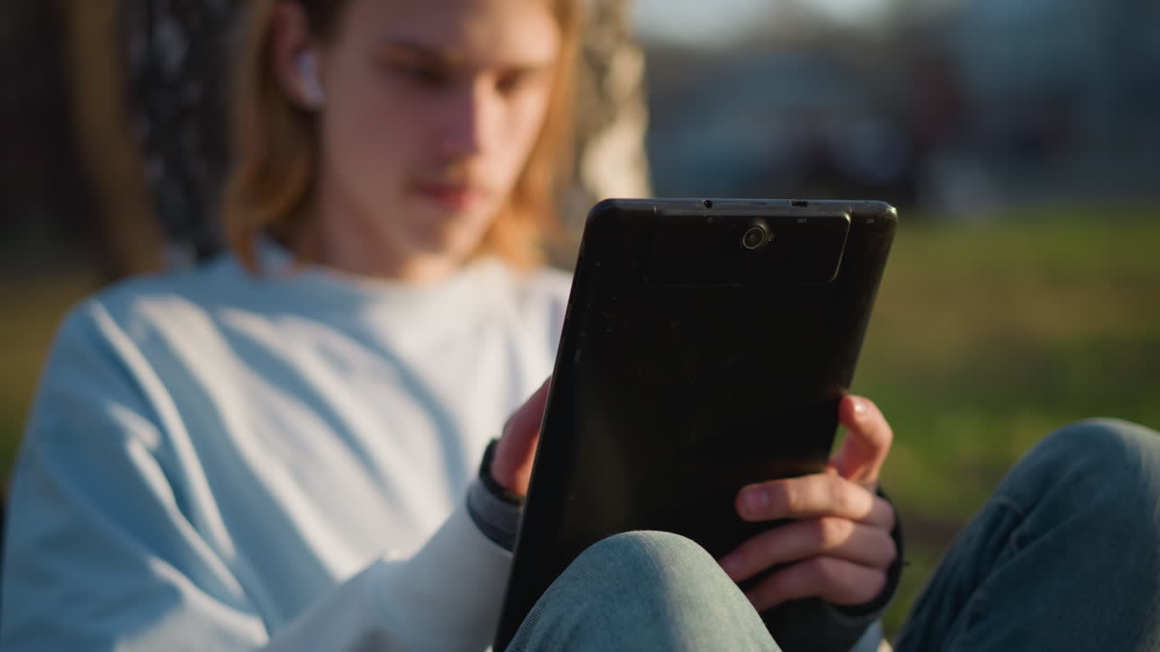 primer plano de un adolescente blanco usando una tableta en un parque, con las manos y los dedos desplazándose por la pantalla con auriculares, sentado en la hierba durante una hora dorada cálida, un estudiante relajado investigando contenido y disfrutando de un suave bokeh