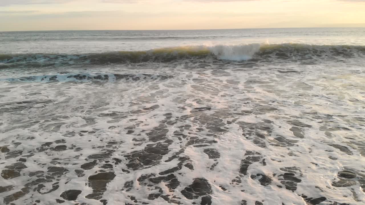 Drone flying forwards and upwards over waves breaking on beach at sunset