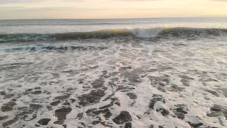 Drone-flying-forwards-and-upwards-over-waves-breaking-on-beach-at-sunset