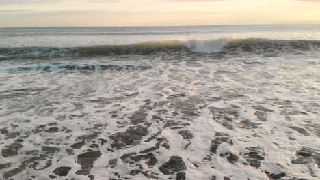 drone flying forwards and upwards over waves breaking on beach at sunset