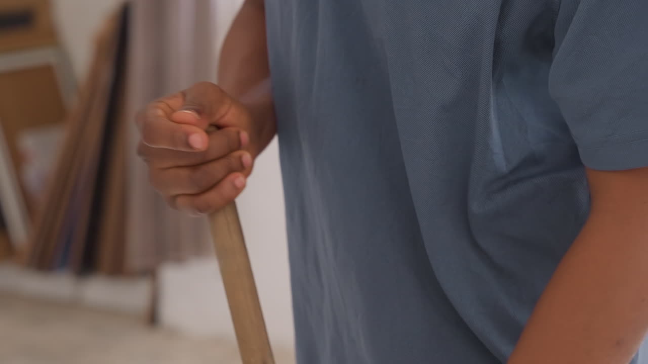 Young person engaged in household cleaning task. Teenager using broom to sweep wooden floor diligently. Junior individual involved in thorough sweeping of wooden floor within domestic setting