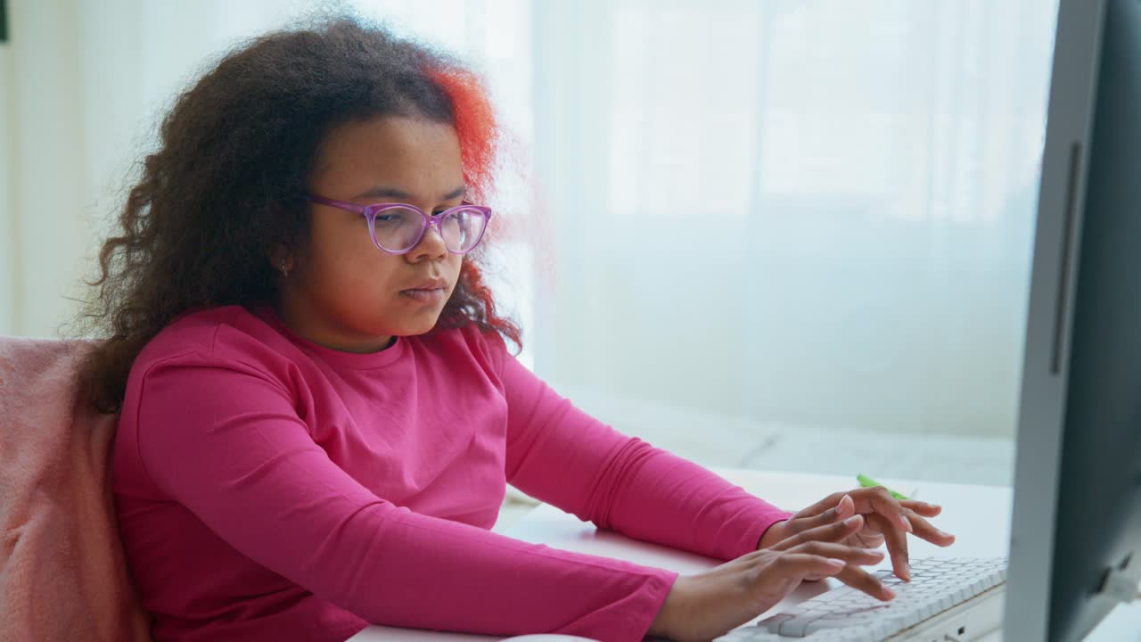 Concentrated young girl with glasses and colorful hair is sitting at a desk, diligently typing on a computer keyboard, focused on her online education