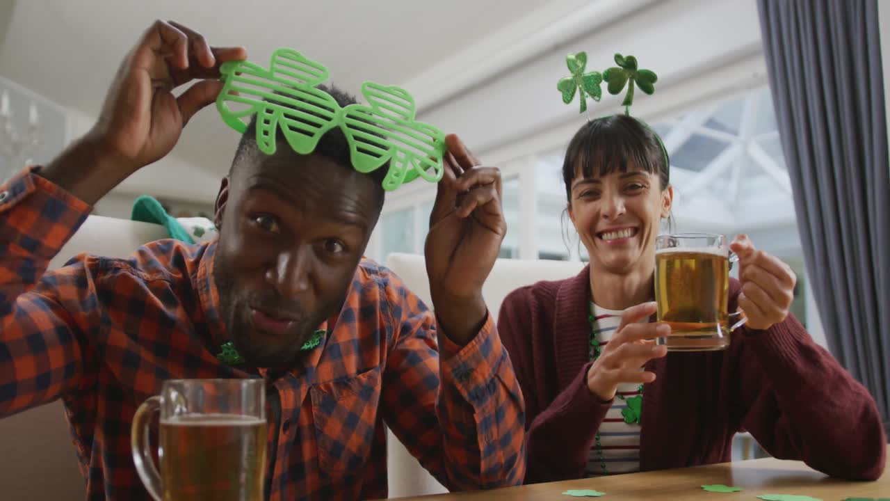 Ritratto di coppia felice e multietnica con birra, che indossa oggetti a forma di trifoglio e sta facendo una videochiamata.
