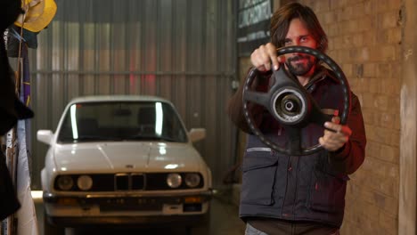 man holds up steering wheel pretending to drive with car in background