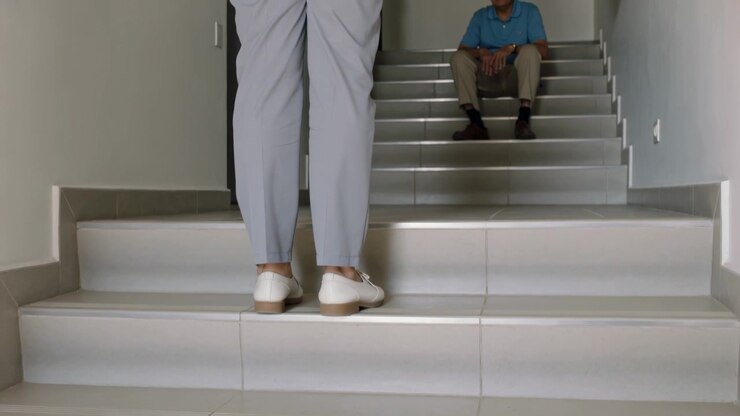 Stepping up riser, adult in gray pants coming to seated senior on tiled stairs, checking welfare