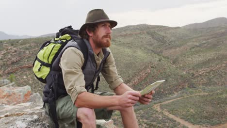 bearded caucasian male survivalist sitting on mountain in wilderness, reading map and looking around