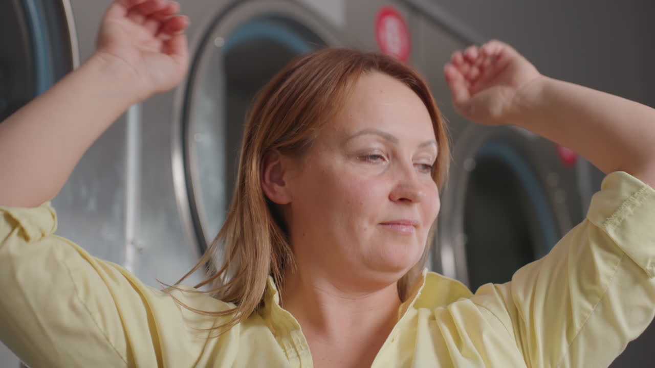 Portrait of female entrepreneur raising hair, arms up before industrial laundry dryers, clothes rolling behind glass, stainless machines, glowing controls, modern laundromat reliability